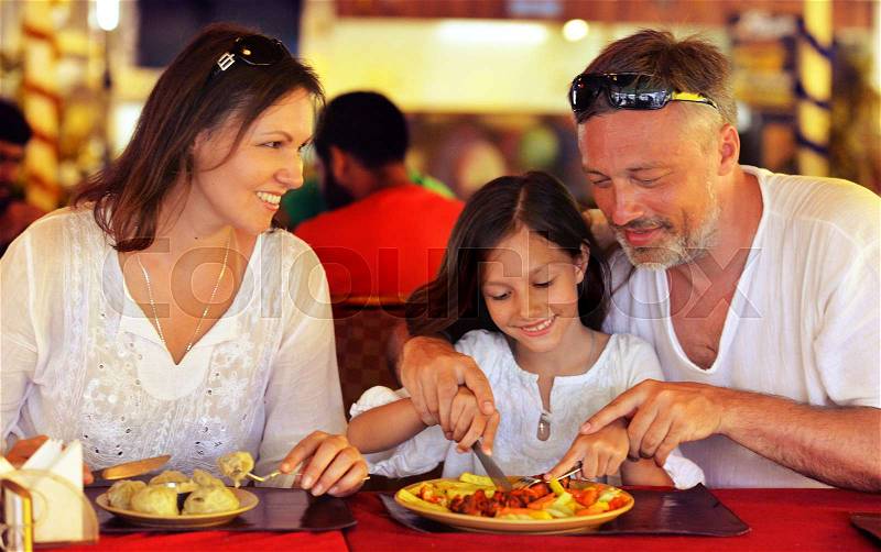 Smiling parents teaching their daughter to use knife in cafe, stock photo