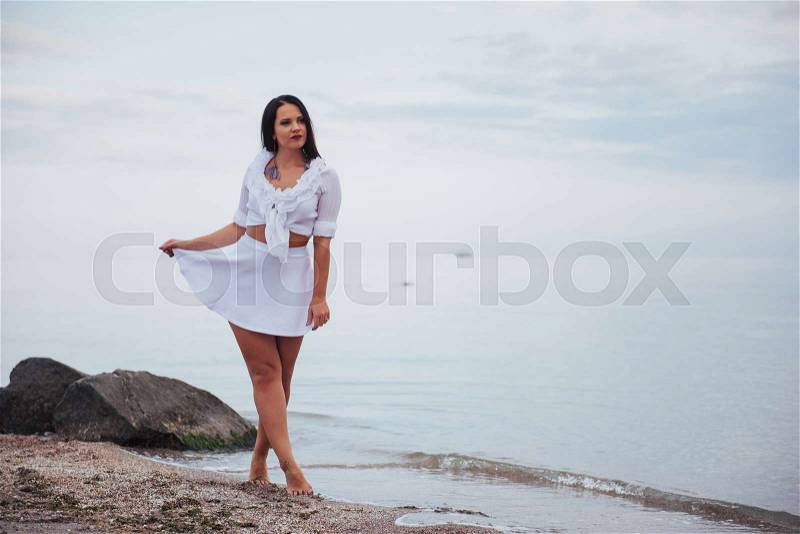 Woman in a white shirt and skirt dancing barefoot on the sand beach hand above the azure sea breeze shakes her long hair, stock photo