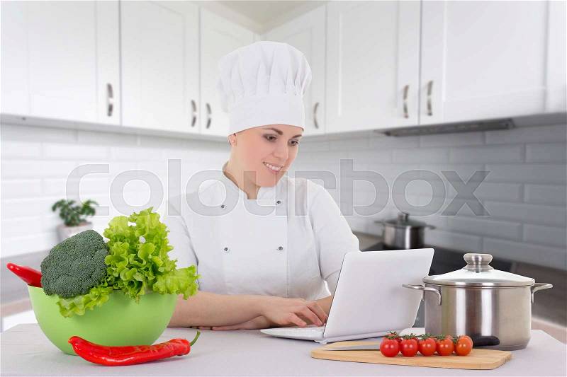 Portrait of attractive cook woman in uniform sitting in the kitchen with laptop, stock photo