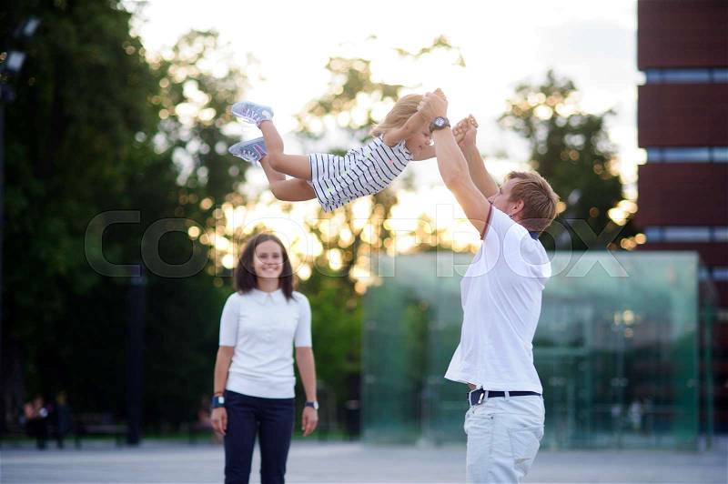 Young family on walk. The father cheerfully plays with the little daughter. He throws up his baby. Mother stands and smiles, stock photo