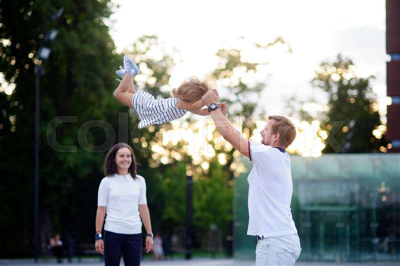Young family on walk. The father cheerfully plays with the little daughter. He throws up his baby. Mother stands and smiles, stock photo