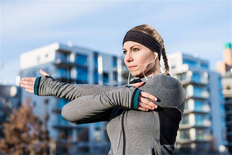 Portrait of a determined young woman stretching her left arm during warm-up routine before outdoor workout in a sunny day, stock photo