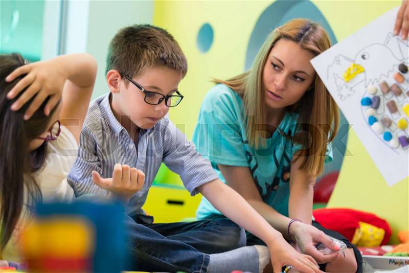 Cute pre-school boy wearing eyeglasses while learning to read from a book for children helped by a dedicated kindergarten teacher assistant, stock photo