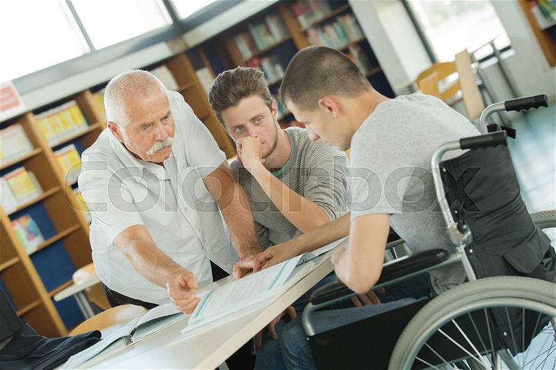 Student in wheelchair talking with classmate and teacher in library, stock photo