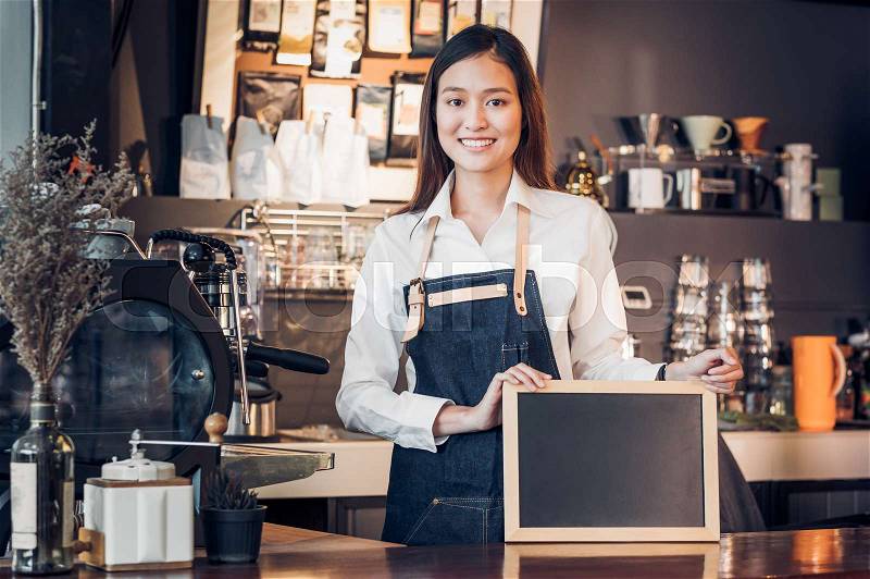 Asian female barista wear jean apron and holding blank blackboard menu at counter bar with smile face,cafe service concept,owner business start up,mock up for display of drink menu or text, stock photo