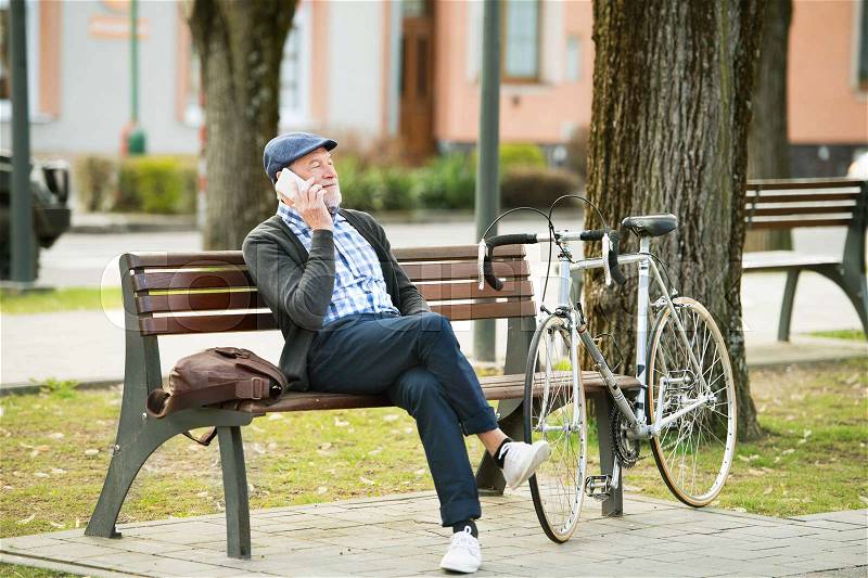 Handsome senior man with bicycle in town park sitting on bench, holding smart phone, making phone call. Sunny spring day, stock photo