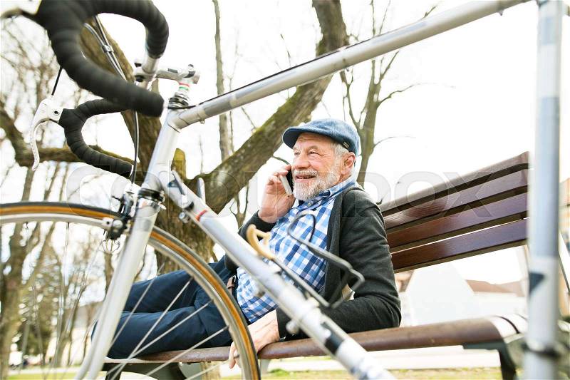 Handsome senior man with bicycle in town park lying on bench, holding smart phone, making phone call. Sunny spring day, stock photo