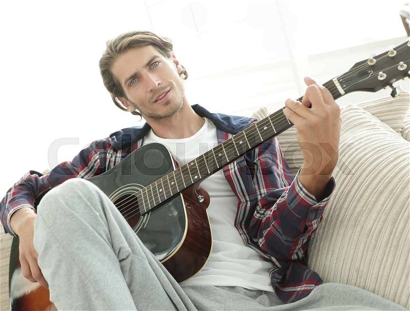 Stylish guy with guitar sitting on sofa in living room, stock photo