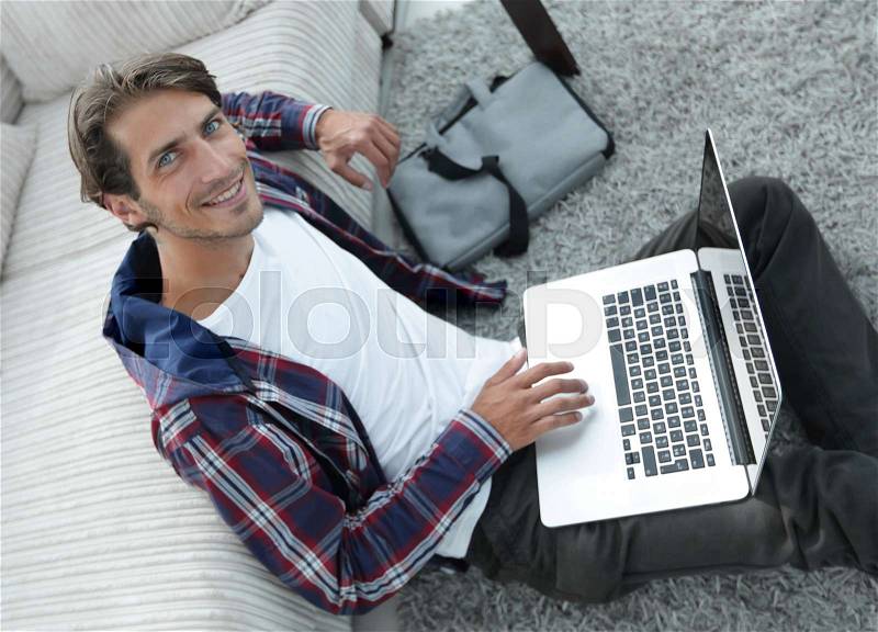 Business guy with laptop sitting near sofa on carpet in living room, stock photo