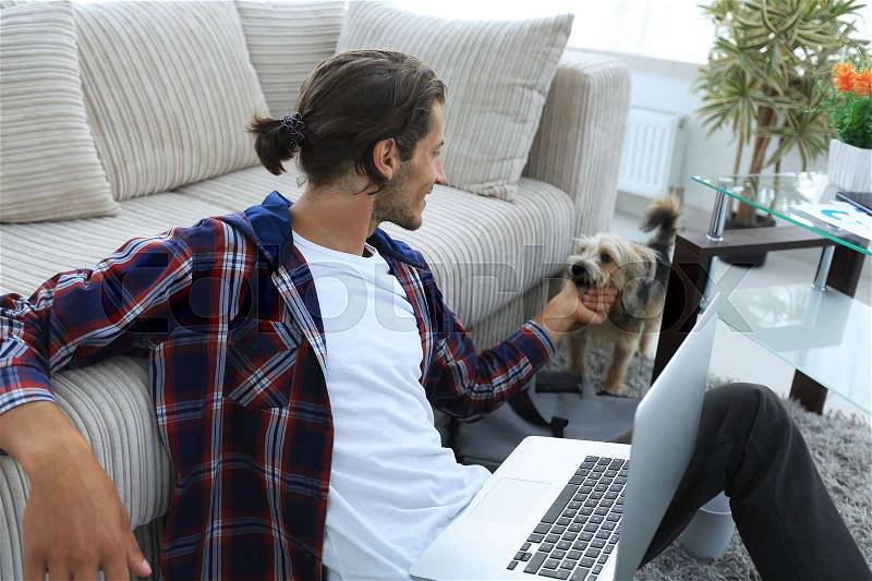 Stylish young man stroking his pet and working on laptop in living room. concept of a lifestyle, stock photo