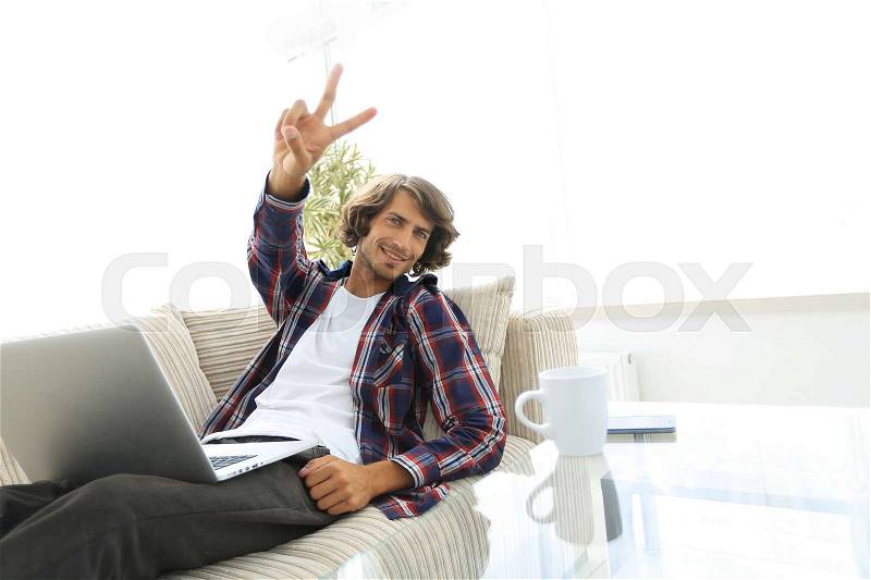 Guy with laptop sitting on sofa and showing hand winning triumph. concept of a lifestyle, stock photo