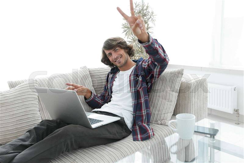 Guy with laptop sitting on sofa and showing hand winning triumph. concept of a lifestyle, stock photo