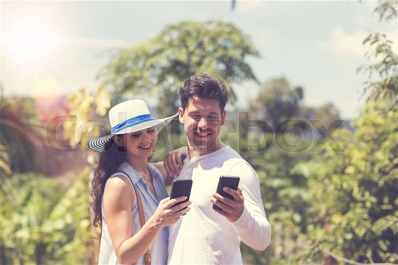 Attractive Couple Messaging Online Using Smart Phones Man And Woman Embracing Over Tropical Forest Landscape Smiling With Smartphones In Hands, stock photo