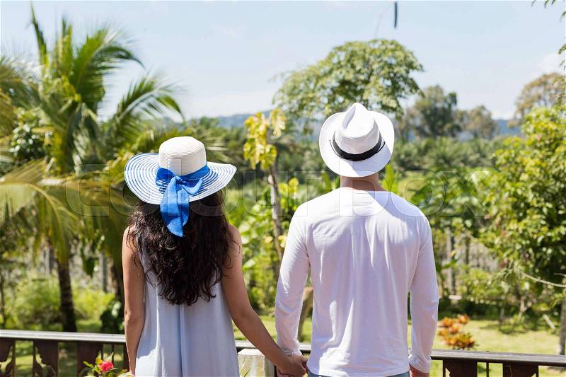 Back Rear View Of Young Couple Wearing Hats Holding Hands Looking At Beautiful Tropical Landscape On Balcony Or Summer Terrace, stock photo