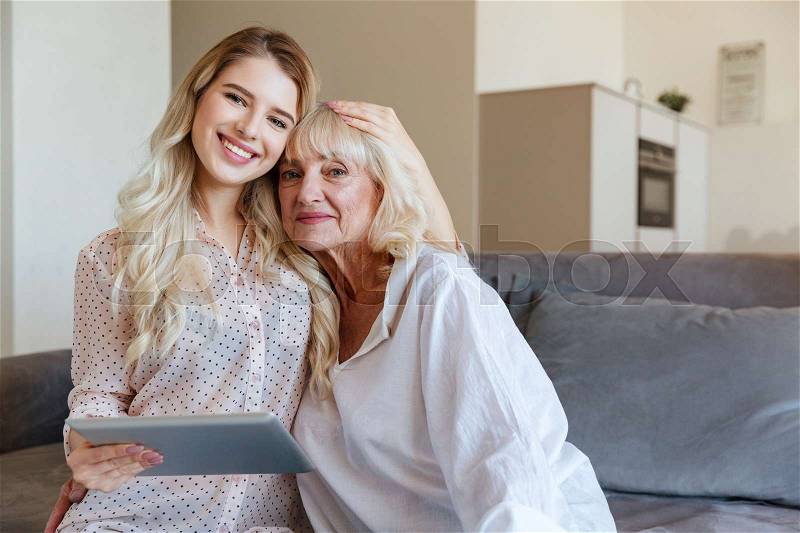 Picture of happy young lady sitting at home with her grandmother using tablet computer. Looking camera, stock photo