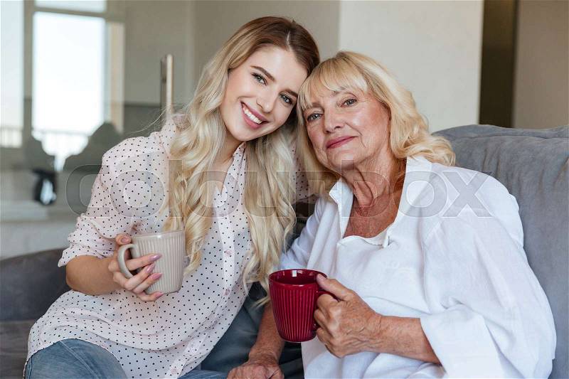 Picture of smiling young lady sitting at home with her grandmother drinking tea. Looking camera, stock photo