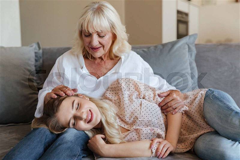 Image of smiling young lady lies on sofa at home with her grandmother hugging. Looking aside, stock photo