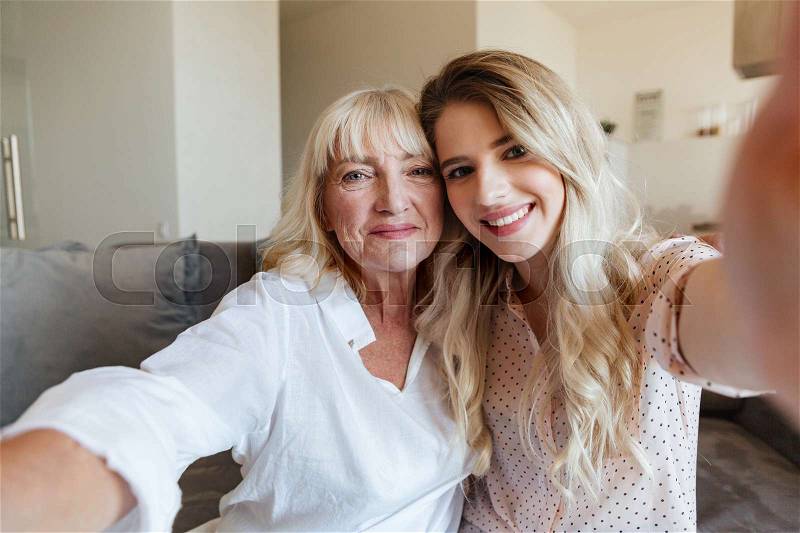 Image of cheerful young lady sitting on sofa at home with her grandmother hugging. Looking camera make selfie, stock photo