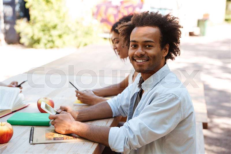Side view of african man sitting by the table outdoors with his friends and looking at the camera, stock photo