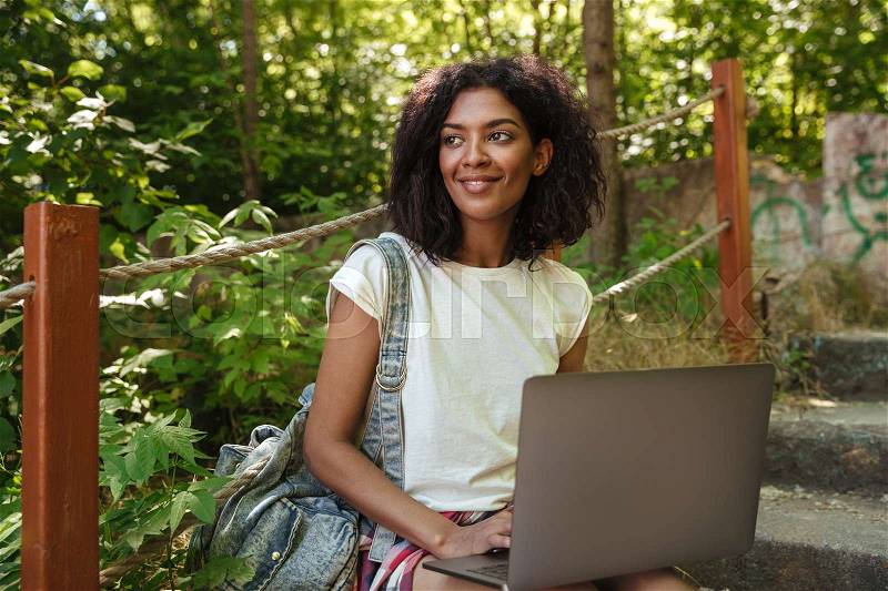 Smiling african woman in forest with laptop computer looking away, stock photo