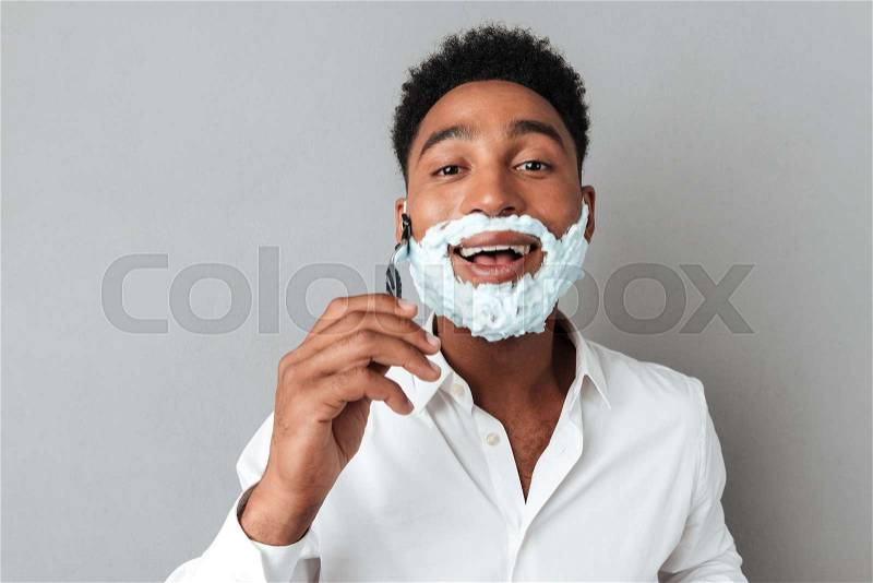 Close up of a young african man in shirt shaving with a razor isolated over gray background, stock photo