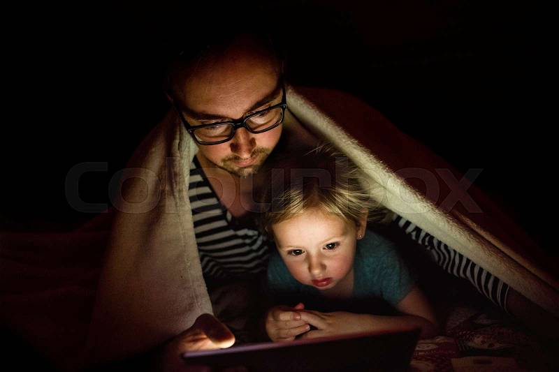 Young father at home with his cute little daughter under blanket at night watching something on tablet, stock photo