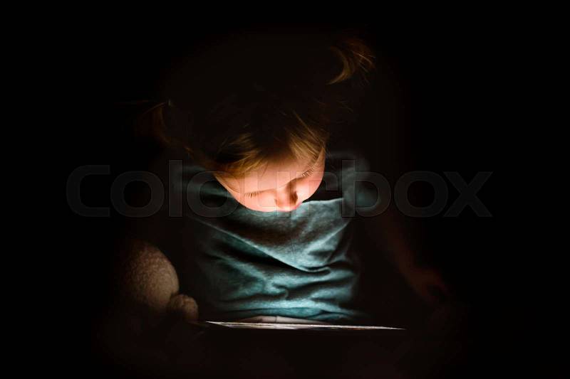 Cute little girl at home at night playing or watching something on tablet, stock photo