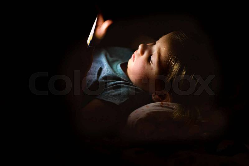 Cute little girl at home at night lying on bed playing or watching something on smart phone, stock photo