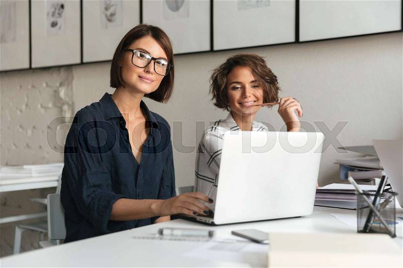 Two young happy women working by the table and looking at the camera in co working office, stock photo