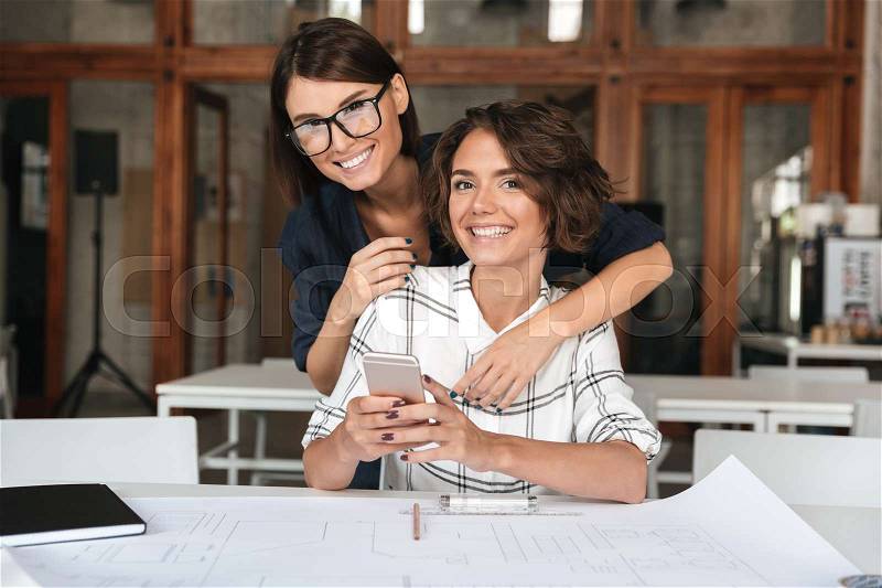 Two young happy women using smartphone by the table in co working office over glass doors background, stock photo