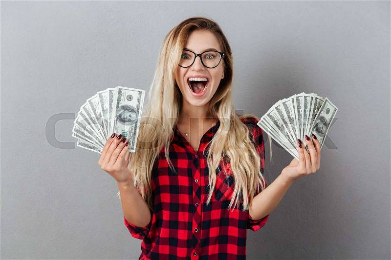 Image of happy young blonde woman wearing glasses standing isolated over grey wall. Looking camera holding money, stock photo