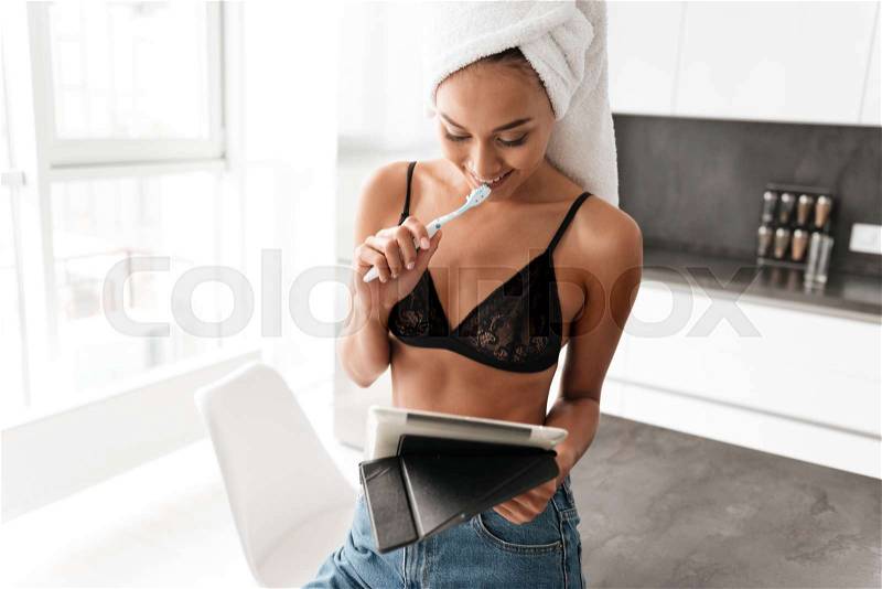 Smiling asian woman with bath towel wrapped around her head brushing her teeth while using tablet computer at the kitchen, stock photo