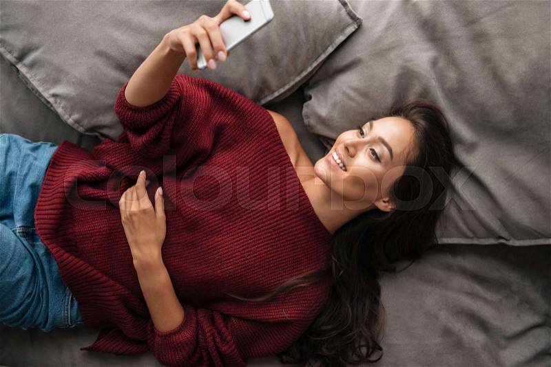Top view of a smiling asian woman in sweater taking a selfie while lying on a couch at home, stock photo