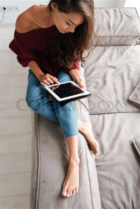 Top view of a young woman in sweater using pc tablet while sitting on a couch at home, stock photo