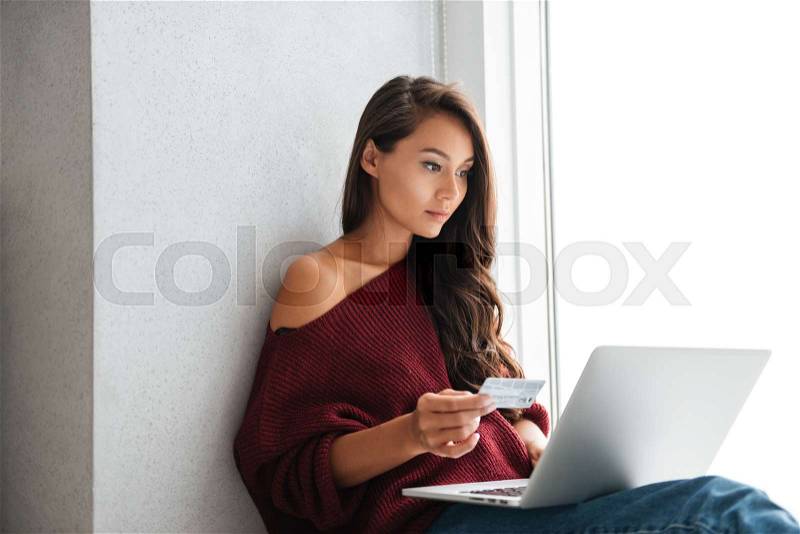 Young beautiful asian woman in sweater holding credit card and looking at laptop screen at home, stock photo