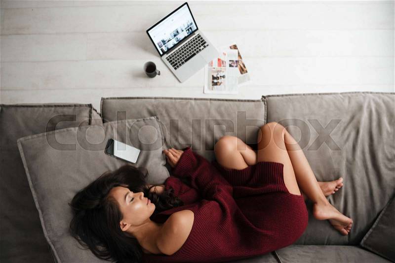 Top view of a beautiful asian woman in sweater sleeping on a couch at home with blank screen mobile phone and laptop, stock photo