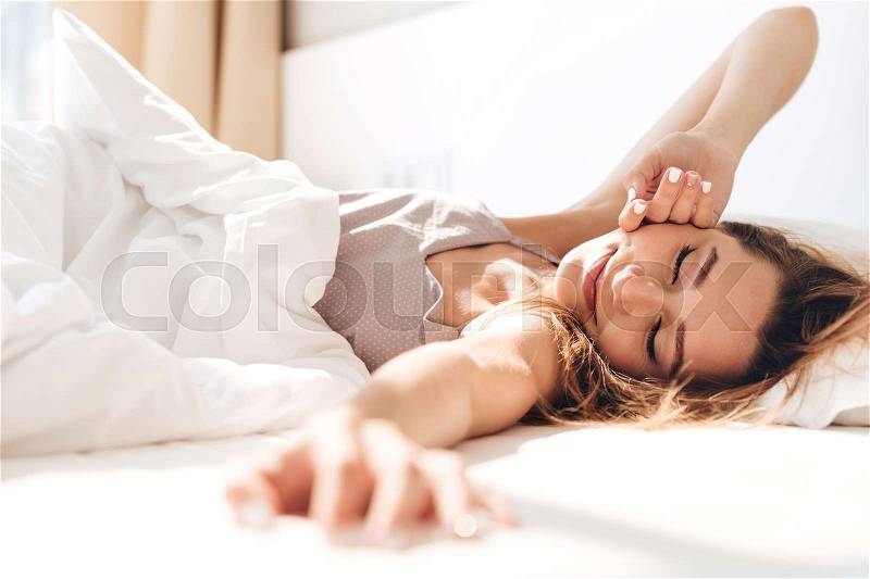 Image of young smiling pretty lady lies in bed indoors. Eyes closed, stock photo