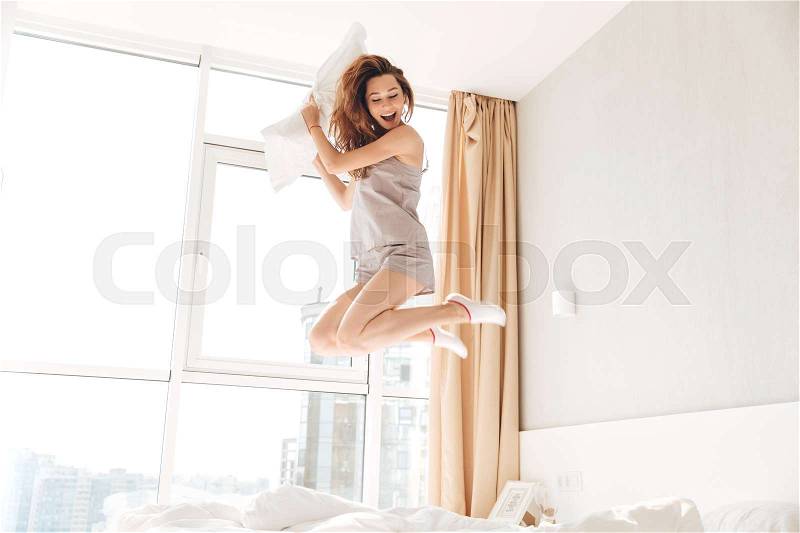 Picture of young happy pretty lady jumping on bed holding pillow indoors. Looking aside, stock photo