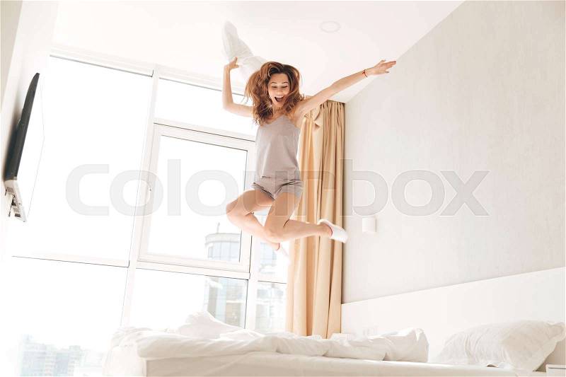 Young funny caucasian woman in pajamas jumping on bed with pillow and smiling at home, stock photo