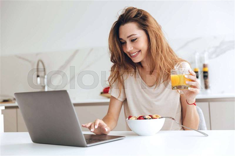 Positive woman with orange juice using laptop computer in the kitchen, stock photo