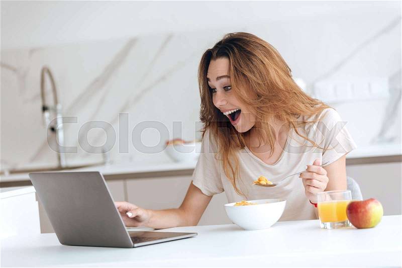 Happy surprised young woman chatting on laptop while have breakfast at home, stock photo