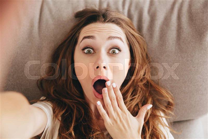 Image of young amazing shocked emotional pretty lady lies on sofa indoors. Looking camera make selfie, stock photo