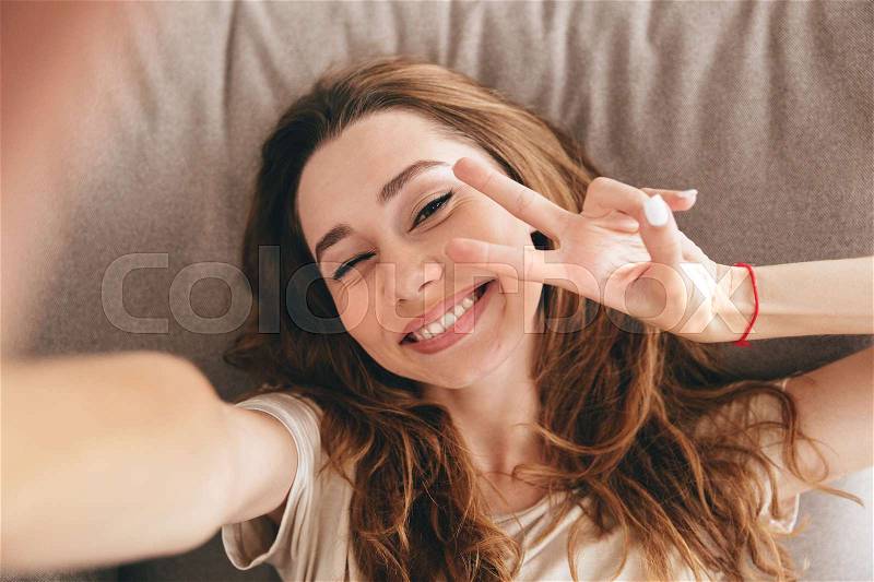 Image of young amazing emotional pretty lady lies on sofa indoors. Looking camera make selfie make peace gesture, stock photo