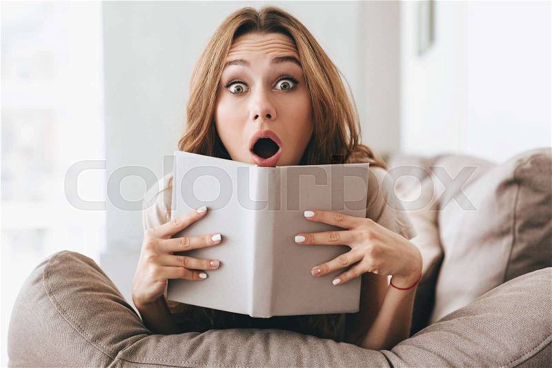Shocked young woman with book sitting on sofa and looking camera at home, stock photo
