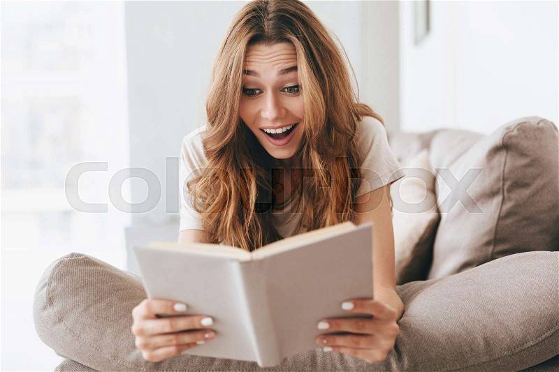 Surprised smiling young woman in casual reading book on sofa at home, stock photo