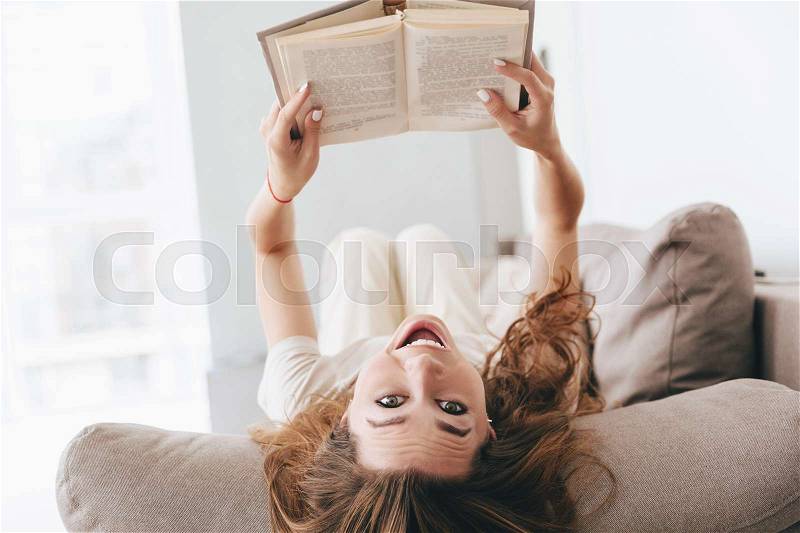 Shot of funny smiling woman lying on couch with book and looking camera at home, stock photo