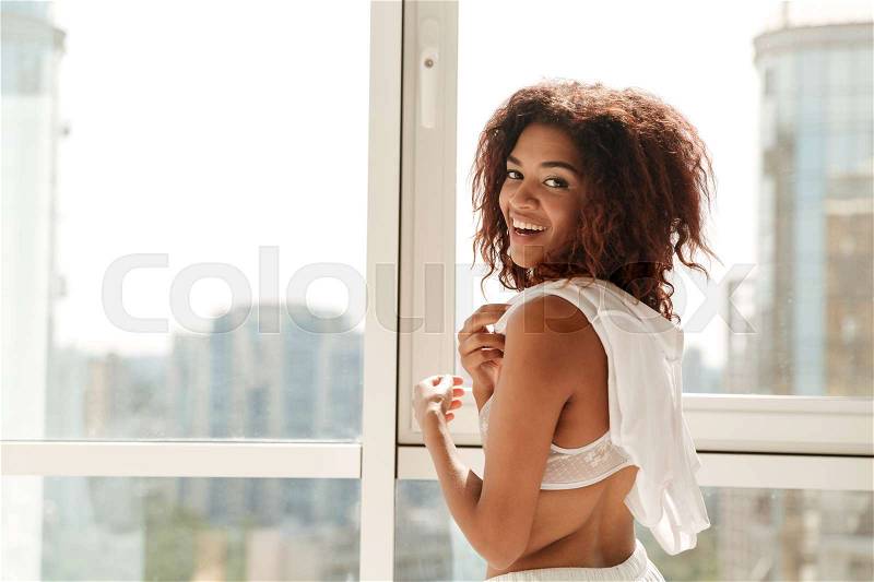 Happy african woman in pajamas laughing near window to camera at home, stock photo