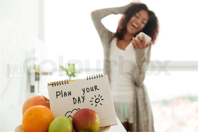 Close up of notebook with slogan \'Plan your day\' between fruits. Laughing lady with smartphone, stock photo