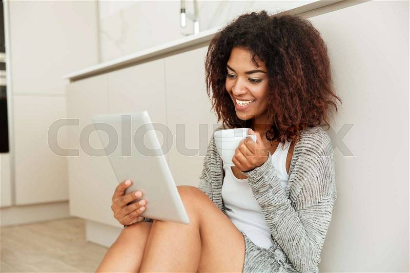 Young cute carefree woman with smile chatting on tablet computer and holding cup of tea, stock photo