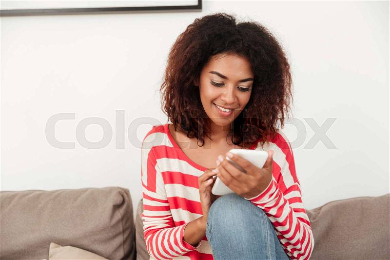 Young african smiling woman on sofa chatting on smartphone while sitting on sofa, stock photo